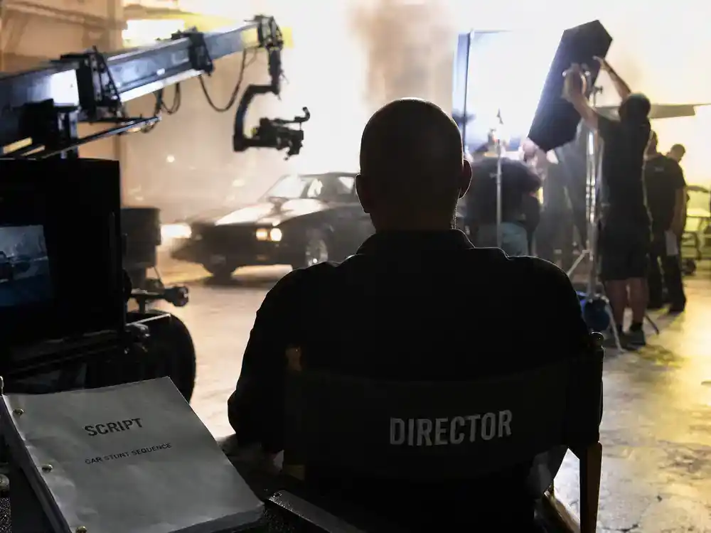 Fast and Furious movies cast order guide Vin Diesel — lead actor silhouette from behind on director chair at car stunt film set with clapperboard