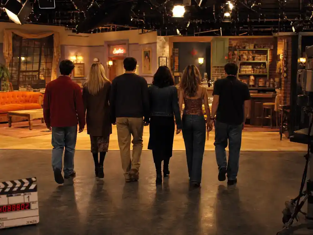 Friends cast net worth 2026 ranked cast production — sitcom coffee shop set corner with film crew and actor silhouettes rehearsing from behind, warm amber lighting