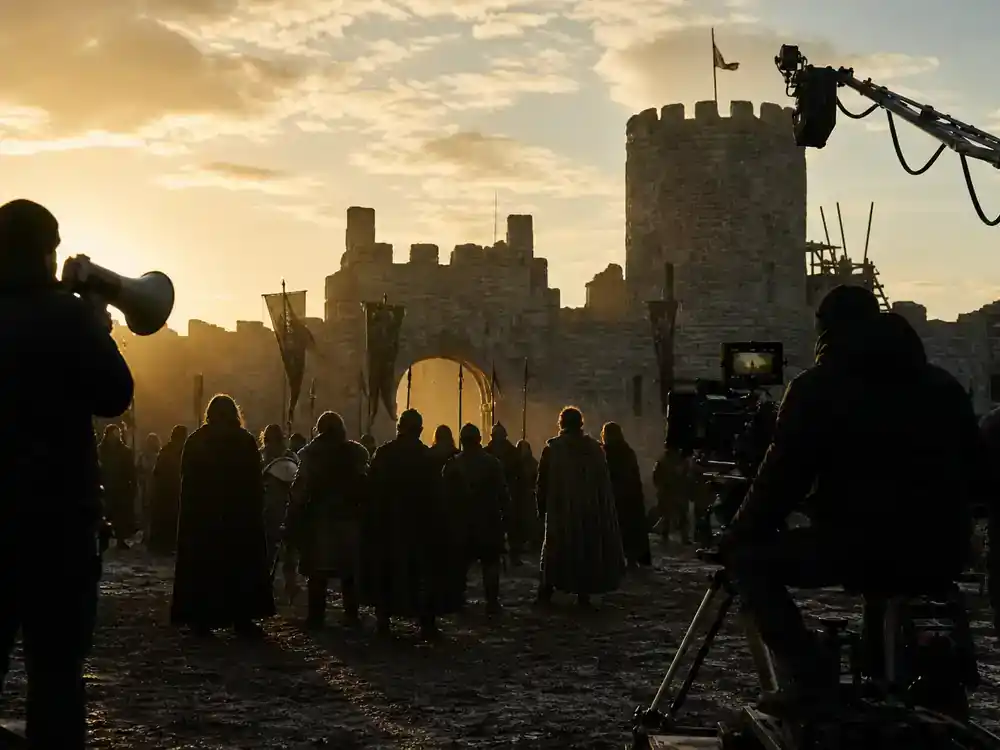 Game of Thrones cast net worth 2026 ranked ensemble — large cast silhouettes on outdoor medieval fantasy TV film location at golden hour with director and camera crew