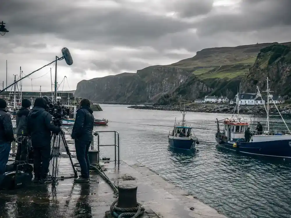Game of Thrones filming locations in Ireland guide Antrim coast — rugged Irish rocky harbor with film crew silhouettes and camera on pier dramatic grey sky