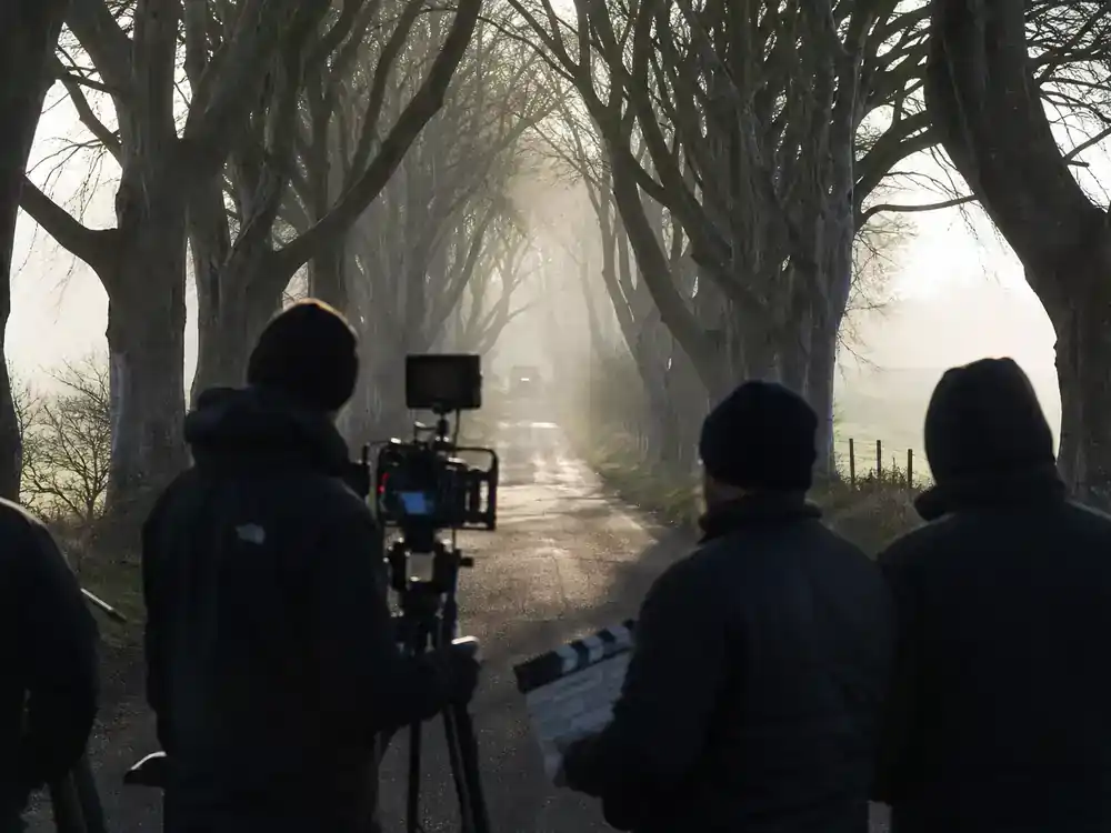Game of Thrones filming locations in Ireland guide Dark Hedges — beech tree tunnel road with film crew silhouettes and camera tripod on Bregagh Road style avenue
