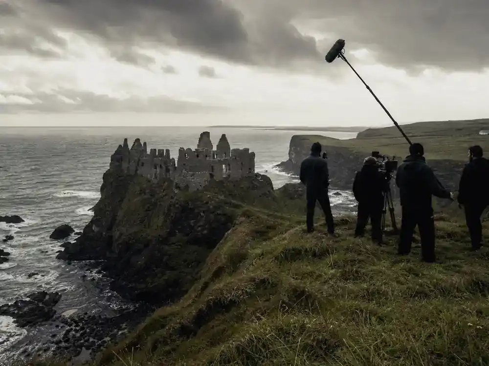 Game of Thrones filming locations in Ireland guide Dunluce Castle — cliffside medieval ruins on Antrim coast with TV drama camera boom and crew silhouettes