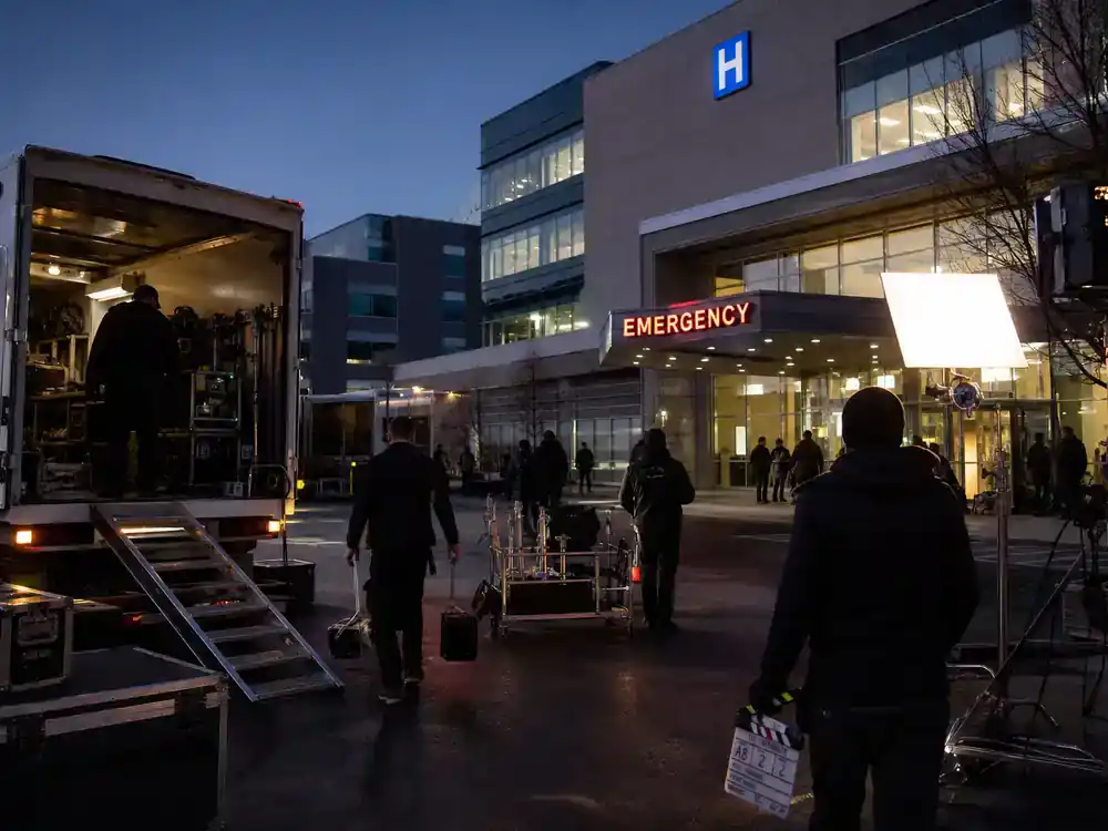 Grey's Anatomy season 22 cast and returning stars hospital filming — film trucks outside modern hospital building with crew silhouettes unloading camera equipment at dusk