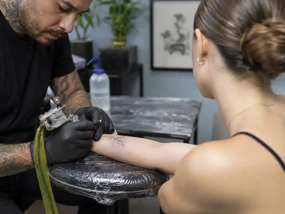 Hailey Bieber tattoos and their meanings studio session — tattoo artist gloved hands working fine-line ink on forearm of client with sleek brown hair from behind