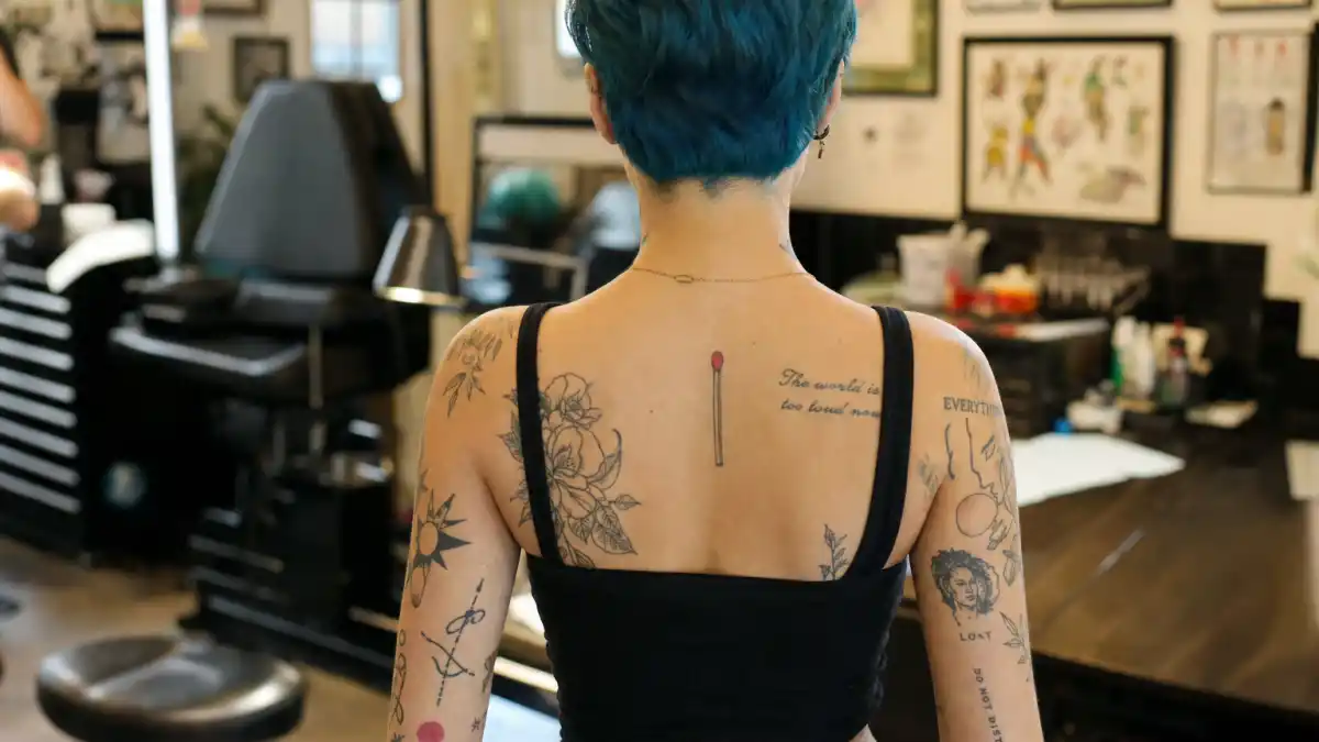 Halsey tattoos and their meanings — pop singer seen from behind in tattoo studio with colorful arm sleeve, matchstick ink, and script tattoos visible