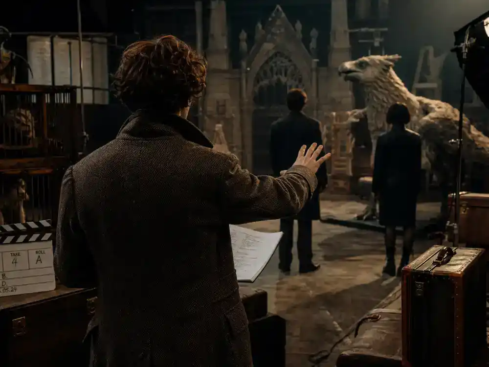 Harry Potter Fantastic Beasts cast guide Newt Scamander — actor silhouette from behind in tweed coat rehearsing on creature-prop soundstage with vintage luggage