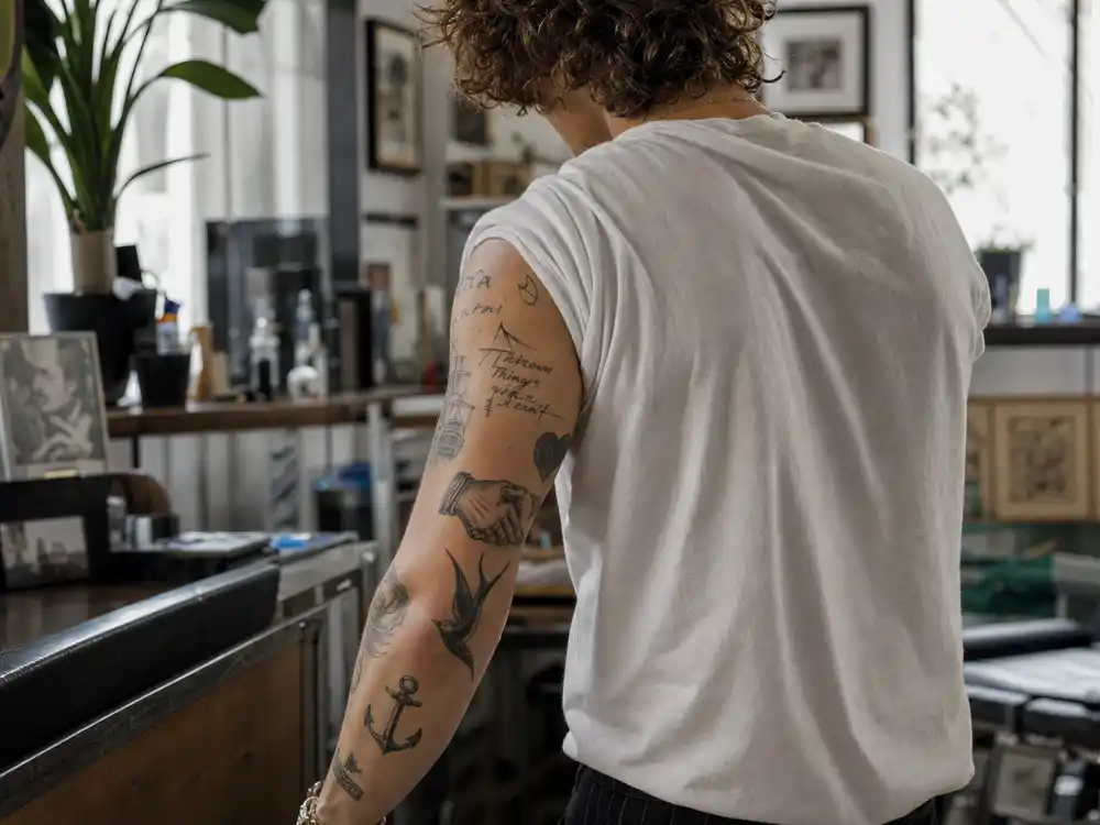 Harry Styles tattoo collection guide arm sleeve — Made in England script cross anchor and swallow bird tattoos on forearm over-shoulder angle