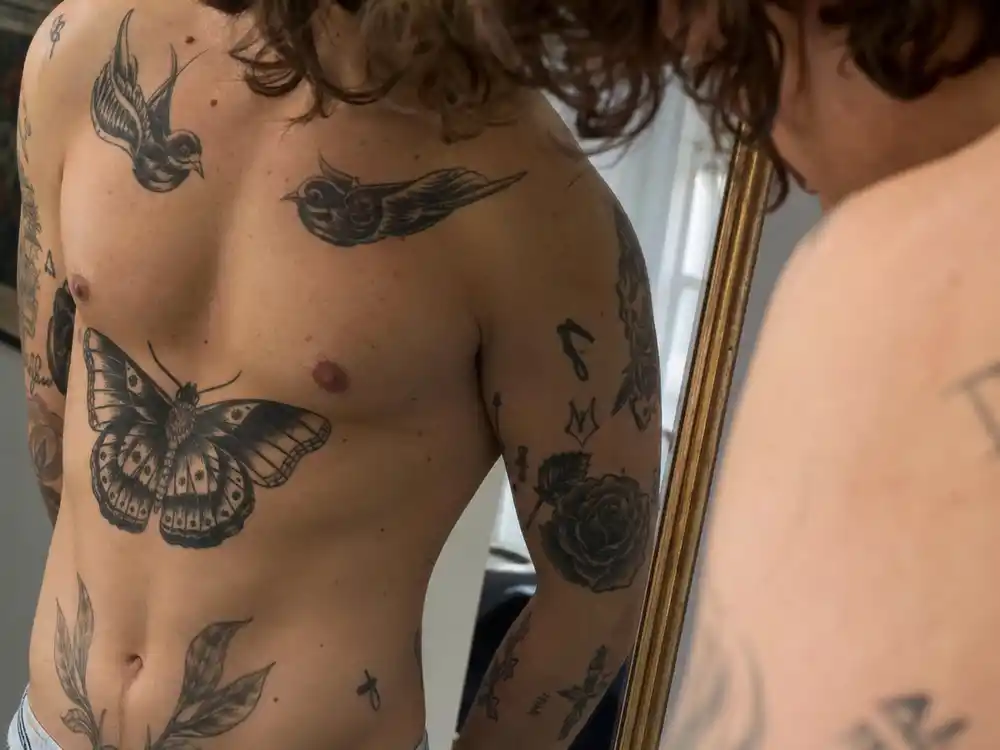 Harry Styles tattoo collection guide butterfly chest — large black butterfly tattoo on tattooed chest and rib floral ink mirror close-up