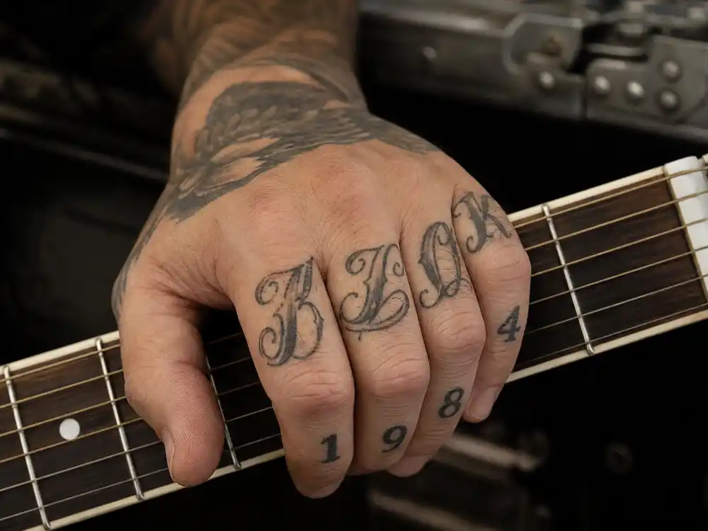 Harry Styles tattoo collection guide hand tattoos — knuckle script numeral and G initial finger ink close-up holding guitar pick