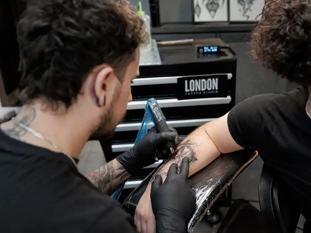 Harry Styles tattoo collection guide studio session — tattoo artist gloved hands applying ink to forearm of curly-haired client over-shoulder angle