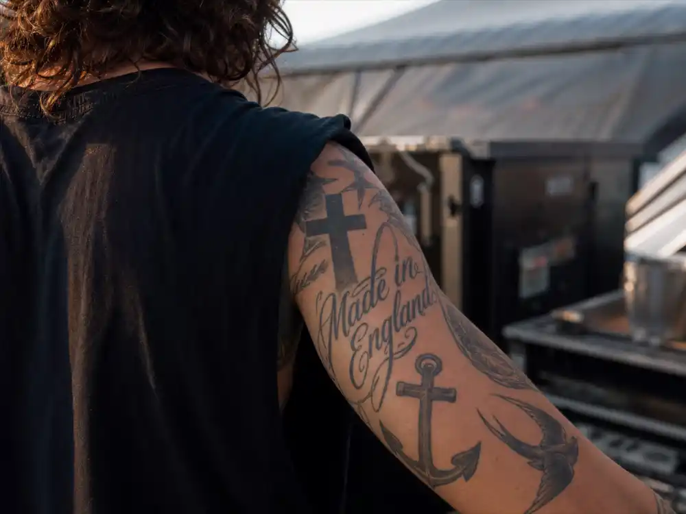 Harry Styles tattoos and their meanings arm sleeve — Made in England script cross anchor and swallow bird tattoos on forearm over-shoulder angle