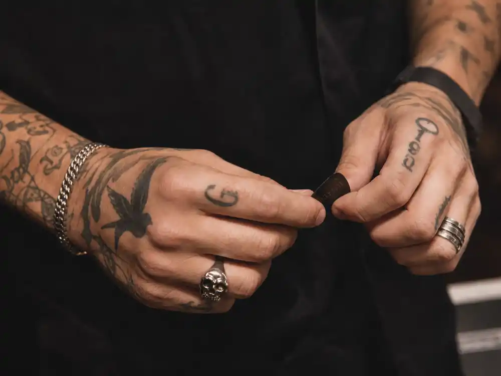 Harry Styles tattoos and their meanings hand tattoos — knuckle script 1970 numeral and G initial finger ink close-up holding guitar pick