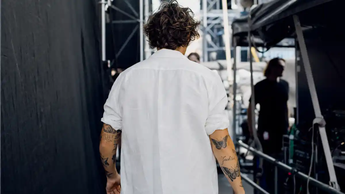 Harry Styles tattoos and their meanings — slim young man with curly brown hair from behind backstage showing butterfly and script arm tattoos