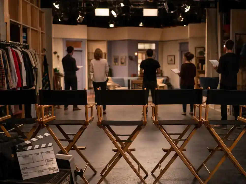 How I Met Your Mother cast net worth 2026 ranked ensemble — five director chairs in a row on TV sitcom soundstage with wardrobe racks and rehearsing actor silhouettes
