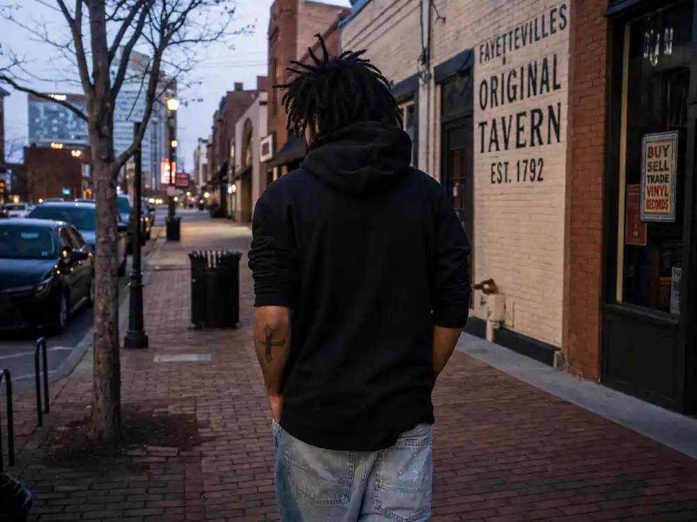 J Cole tattoos and their meanings — artist walking city sidewalk at dusk from behind with cross tattoo peeking from pushed-up sleeve