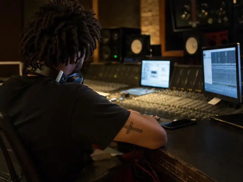 J Cole tattoos and their meanings — recording studio over-shoulder session with cross tattoo on left bicep resting near mixing console