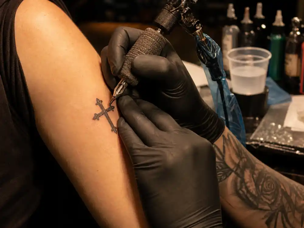 J Cole tattoos and their meanings — professional tattoo studio session with gloved artist applying fine cross design on client upper arm