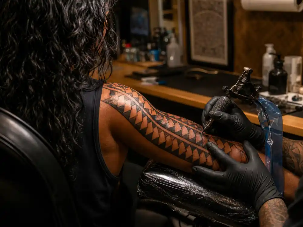 Jason Momoa tattoos and their meanings studio session — tattoo artist gloved hands applying shark teeth triangle pattern on forearm over-shoulder angle