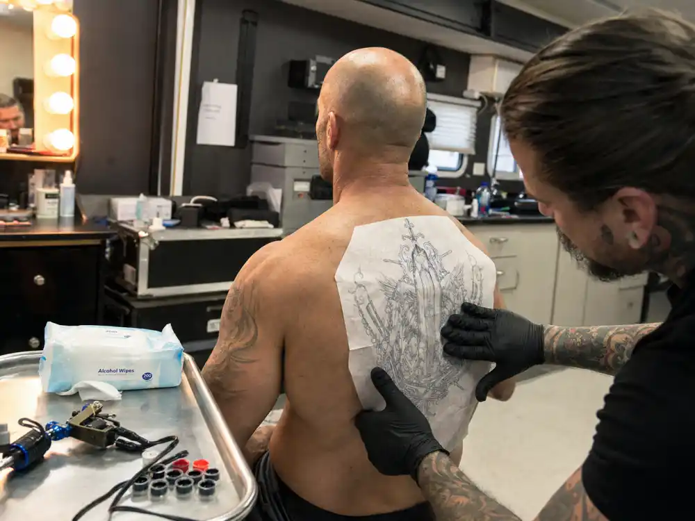 Jason Statham tattoos and their meanings — film makeup artist applying temporary tattoo transfers to action star back on set