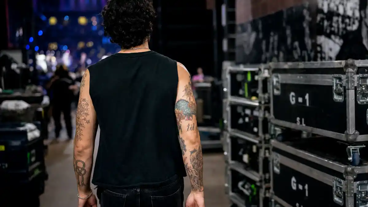 Joe Jonas tattoos and their meanings — dark curly-haired musician from behind backstage showing rope knot and script forearm tattoos