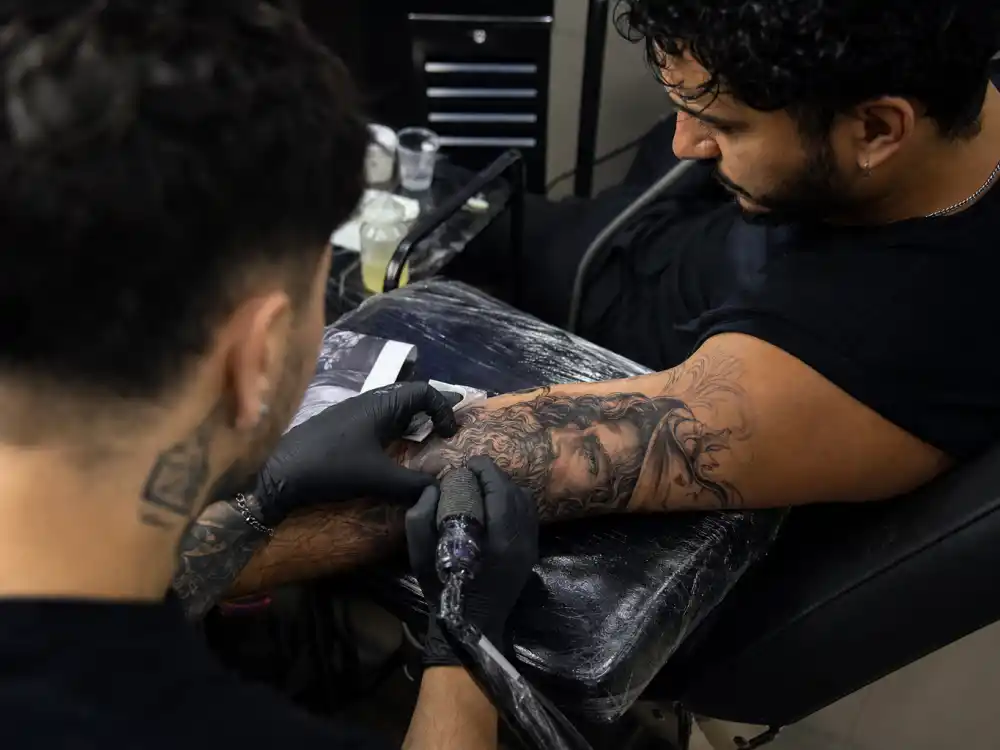Joe Jonas tattoos and their meanings studio session — tattoo artist gloved hands applying portrait ink to dark curly-haired client forearm