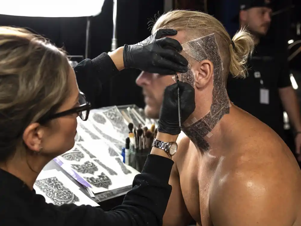 John Cena tattoos and their meanings — makeup artist applying temporary facial tattoo prosthetics on film set for acting role
