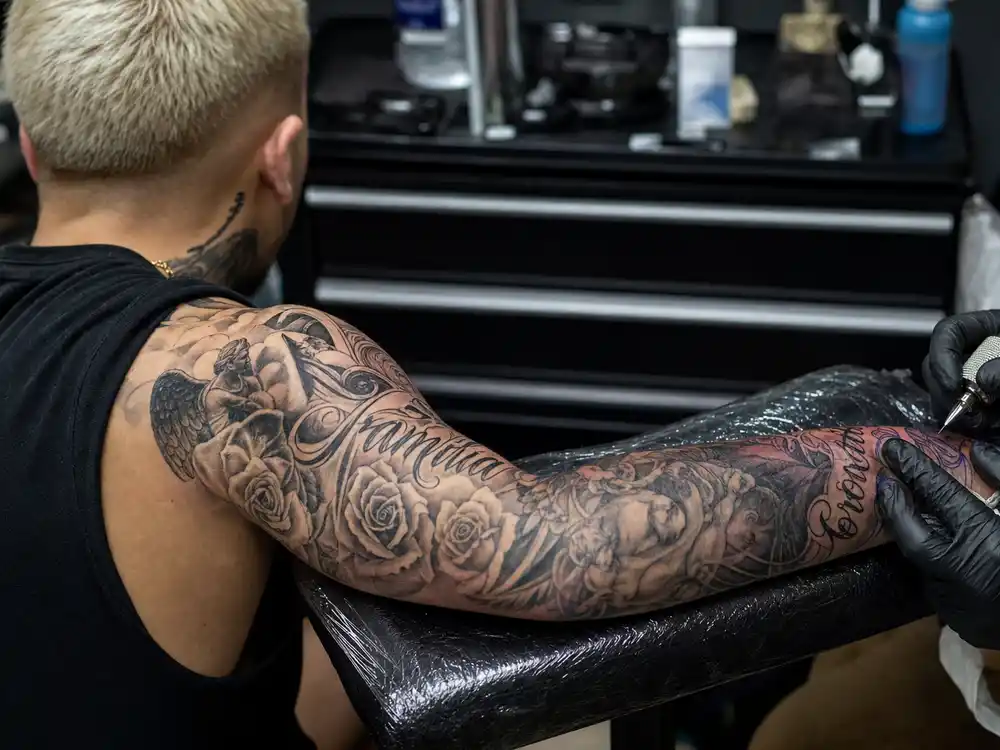 Justin Bieber tattoo collection guide arm sleeve — roses angels and script lettering covering forearm and bicep of young male with bleached blonde hair in tattoo studio