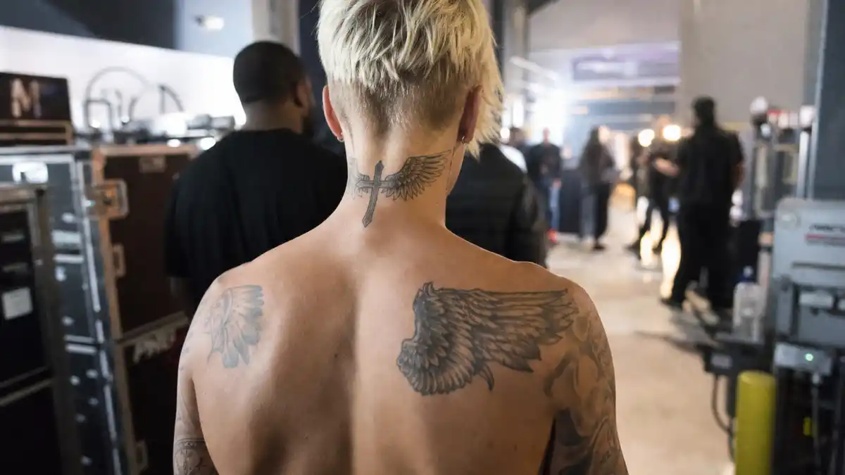 Justin Bieber tattoo collection guide — young pop star with bleached blonde hair from behind showing cross neck tattoo and angel wing back ink backstage at concert