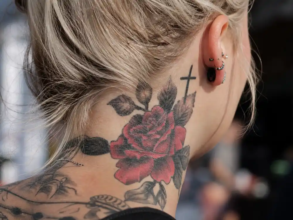 Justin Bieber tattoo collection guide neck rose — rose tattoo on side of neck and cross behind ear on young male with bleached blonde hair over-shoulder view