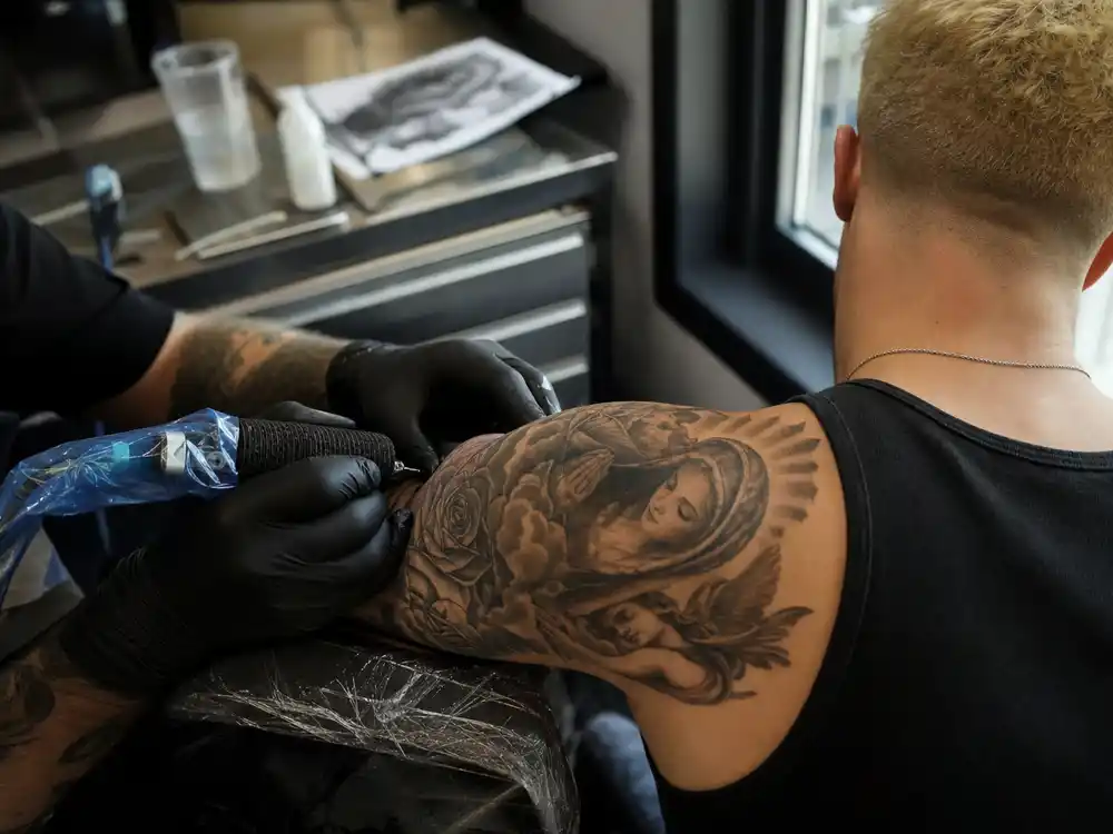 Justin Bieber tattoo collection guide studio session — tattoo artist gloved hands working on religious ink sleeve of young male client with bleached blonde hair from behind