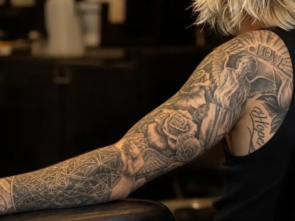 Justin Bieber tattoos and their meanings arm sleeve — roses angels and script lettering covering forearm and bicep of young male with bleached blonde hair in tattoo studio