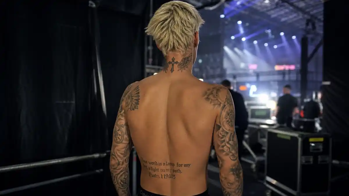 Justin Bieber tattoos and their meanings — young pop star with bleached blonde hair from behind showing cross neck tattoo and full back sleeve ink backstage at concert