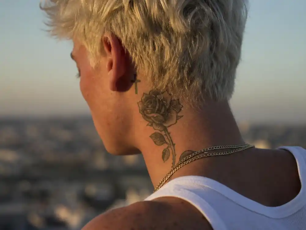 Justin Bieber tattoos and their meanings neck rose — rose tattoo on side of neck and cross behind ear on young male with bleached blonde hair over-shoulder view