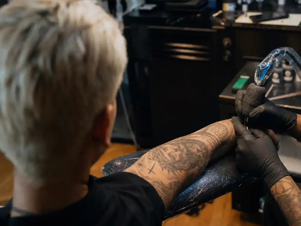 Justin Bieber tattoos and their meanings studio session — tattoo artist gloved hands working on religious ink sleeve of young male client with bleached blonde hair from behind