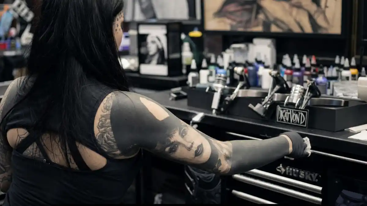 Kat Von D tattoos and their meanings — gothic tattoo artist seen from behind in studio with portrait forearm ink, blackout panels, and star temple tattoos visible