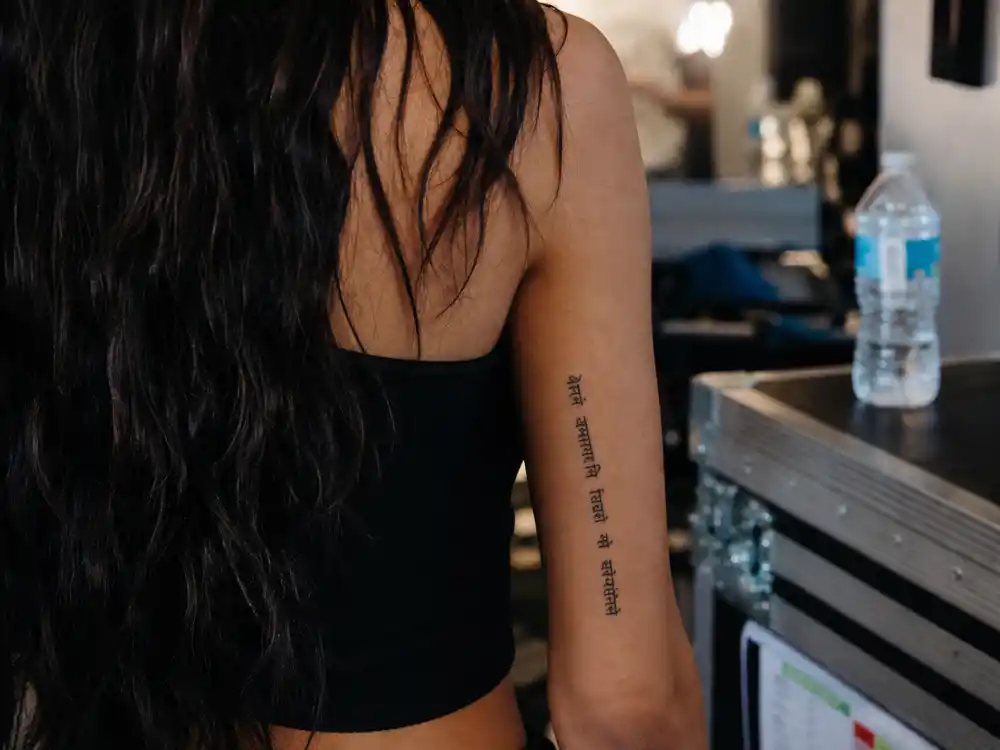 Katy Perry tattoos and their meanings — Sanskrit go with the flow script tattoo on inside of right bicep over-shoulder view
