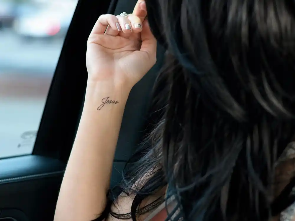 Katy Perry tattoos and their meanings — close-up of Jesus script tattoo on left wrist in fine black lettering