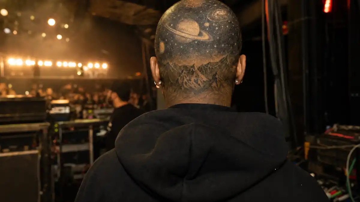 Kid Cudi tattoos and their meanings — hip-hop artist from behind with galaxy scalp tattoo visible under buzz cut at backstage concert