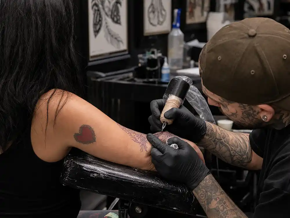 Kylie Jenner tattoos and their meanings — tattoo artist gloved hands inking fine-line red work on forearm during professional studio session