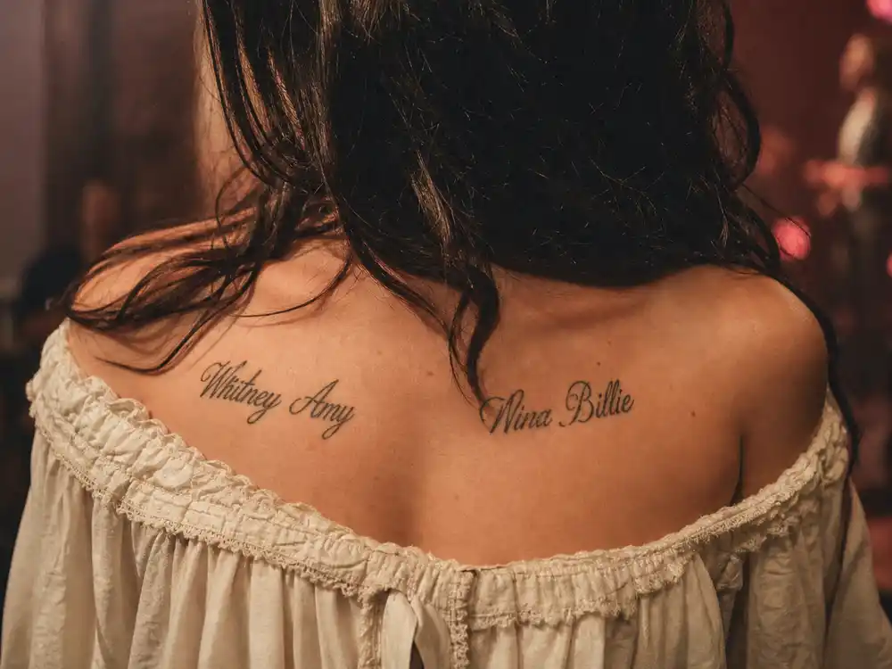 Lana Del Rey tattoos and their meanings chest tributes — Whitney Amy and Nina Billie script tattoos across chest over-shoulder at intimate venue