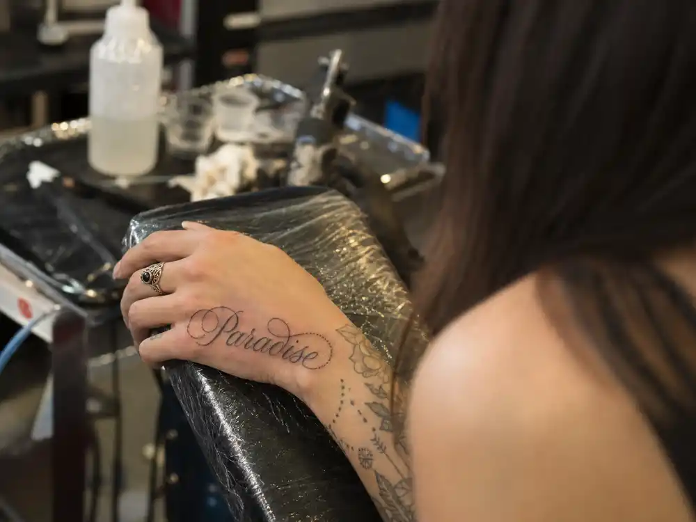 Lana Del Rey tattoos and their meanings Paradise — cursive Paradise script tattoo on left hand close-up at tattoo studio vanity