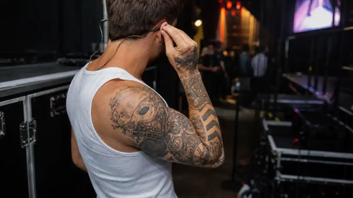 Liam Payne tattoos and their meanings — young British musician with brown hair from behind backstage showing chevron arrow and skull forearm sleeve tattoos