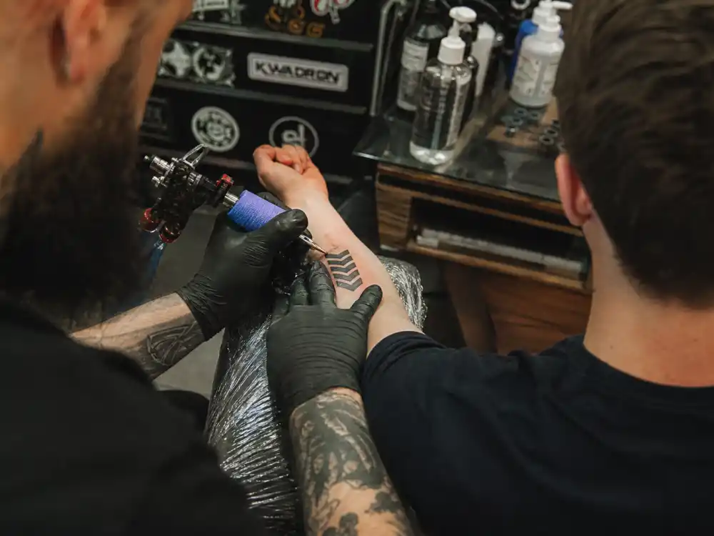 Liam Payne tattoos and their meanings studio session — tattoo artist gloved hands applying chevron arrow ink to brown-haired client forearm over-shoulder angle