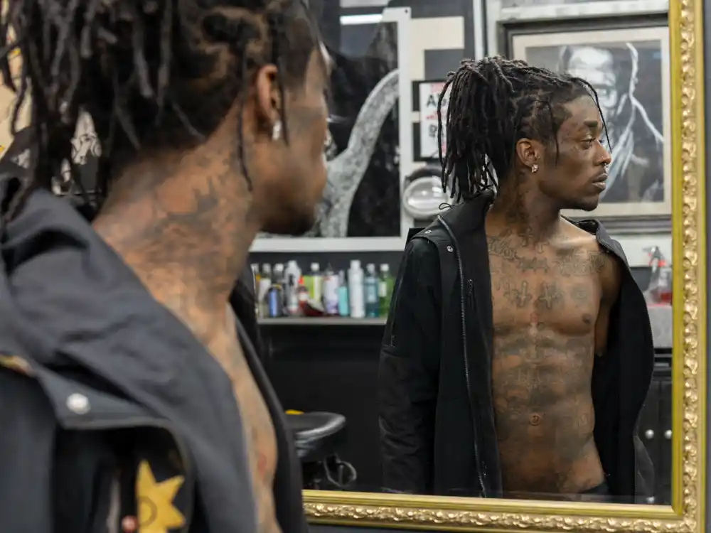 Lil Uzi Vert tattoos and their meanings — chest and neck cross and 444 number tattoos visible over-shoulder on rapper in tattoo shop mirror