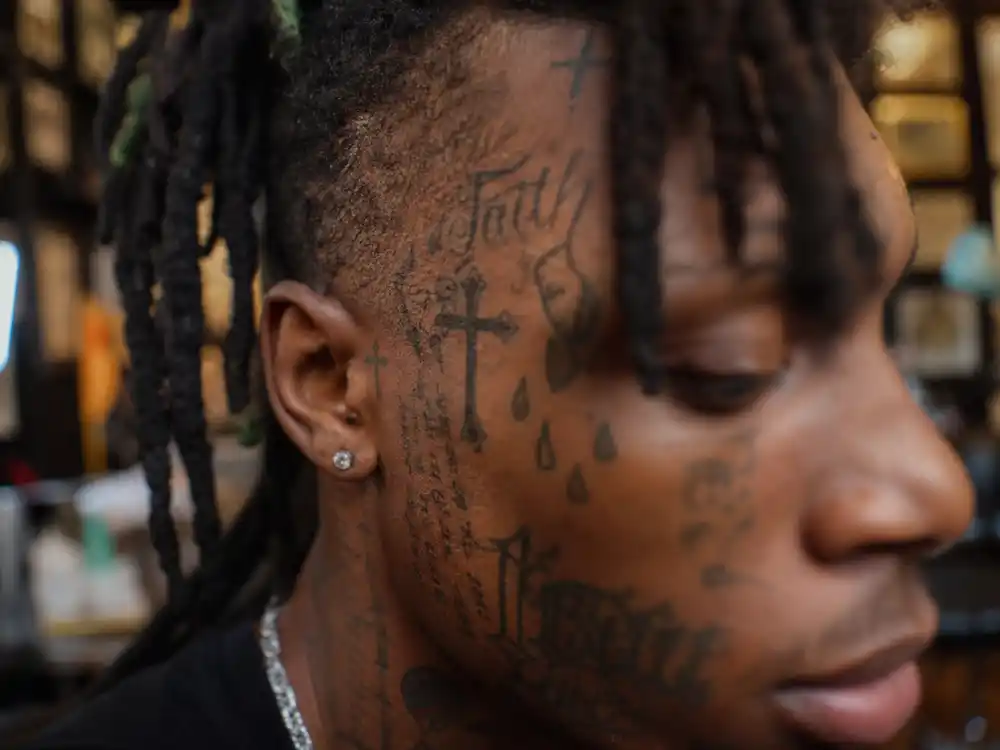 Lil Uzi Vert tattoos and their meanings — close-up of face tattoos including crosses and script lettering on rapper from blurred side angle
