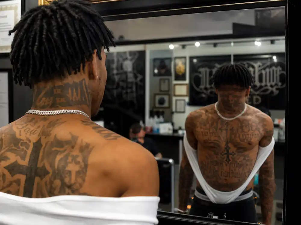 Lil Wayne tattoos and their meanings — chest and neck cross and script tattoos visible over-shoulder on rapper in tattoo shop mirror
