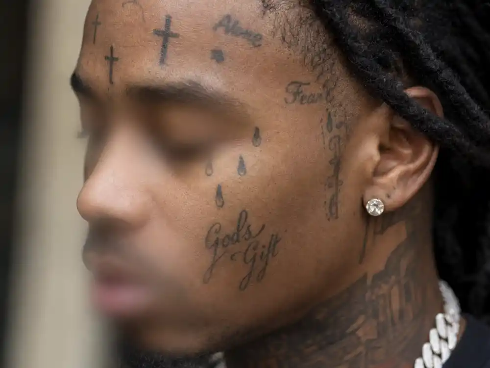 Lil Wayne tattoos and their meanings — close-up of face tattoos including teardrops and cross symbols on rapper from blurred side angle