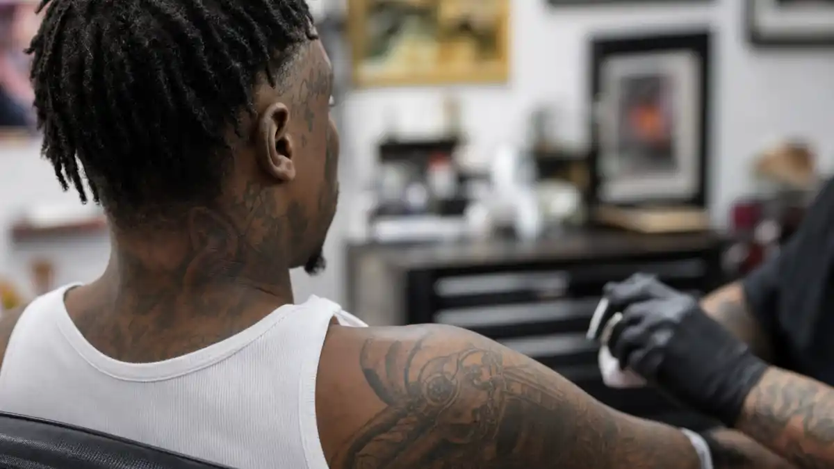 Lil Wayne tattoos and their meanings — rapper seen from behind in tattoo studio with extensive face and sleeve ink visible over-shoulder