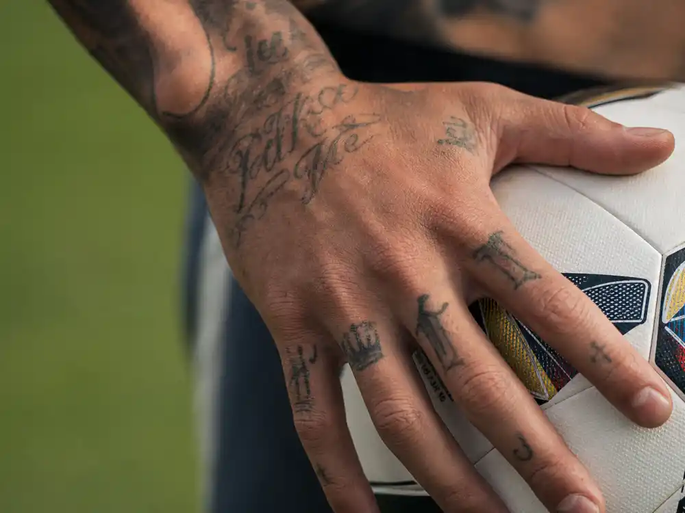 Lionel Messi tattoos and their meanings hand tattoos — crown and script finger ink close-up holding football on pitch