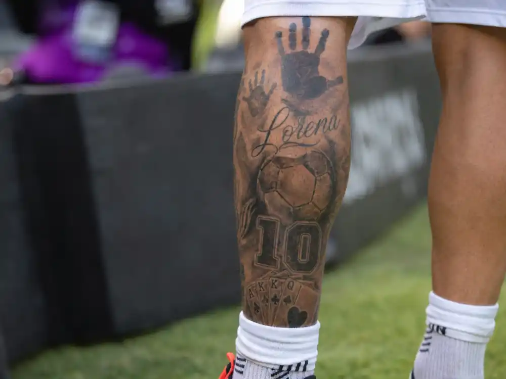 Lionel Messi tattoos and their meanings leg tattoos — left calf close-up showing handprints, number 10, soccer ball and black ink cover-up section